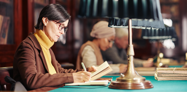A woman takes notes in a notebook while reading at a table. Blurred background features more individuals working at this desk.