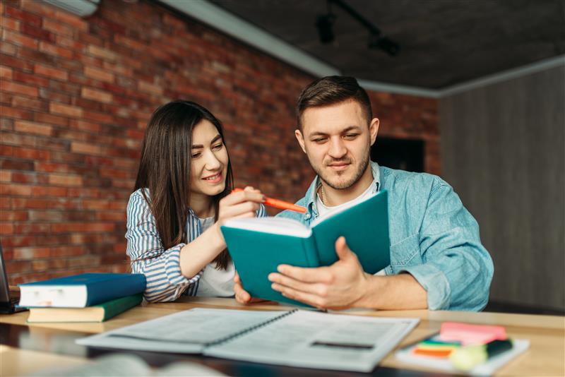 Two individuals looking at and reading something from a textbook together at a table.