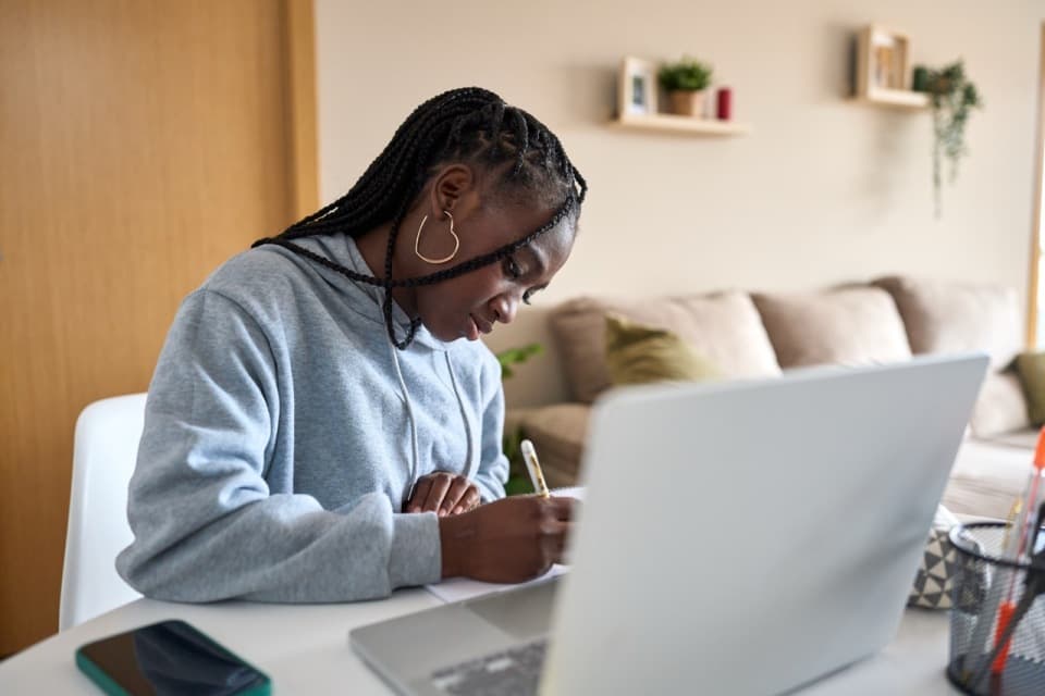 A young woman sits at her desk with her laptop open, taking notes.